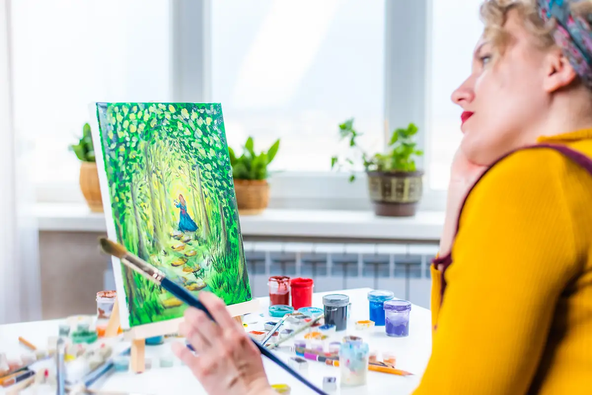 A lady is painting on a canvas while standing back and admiring her work, she is wearing a yellow sweater