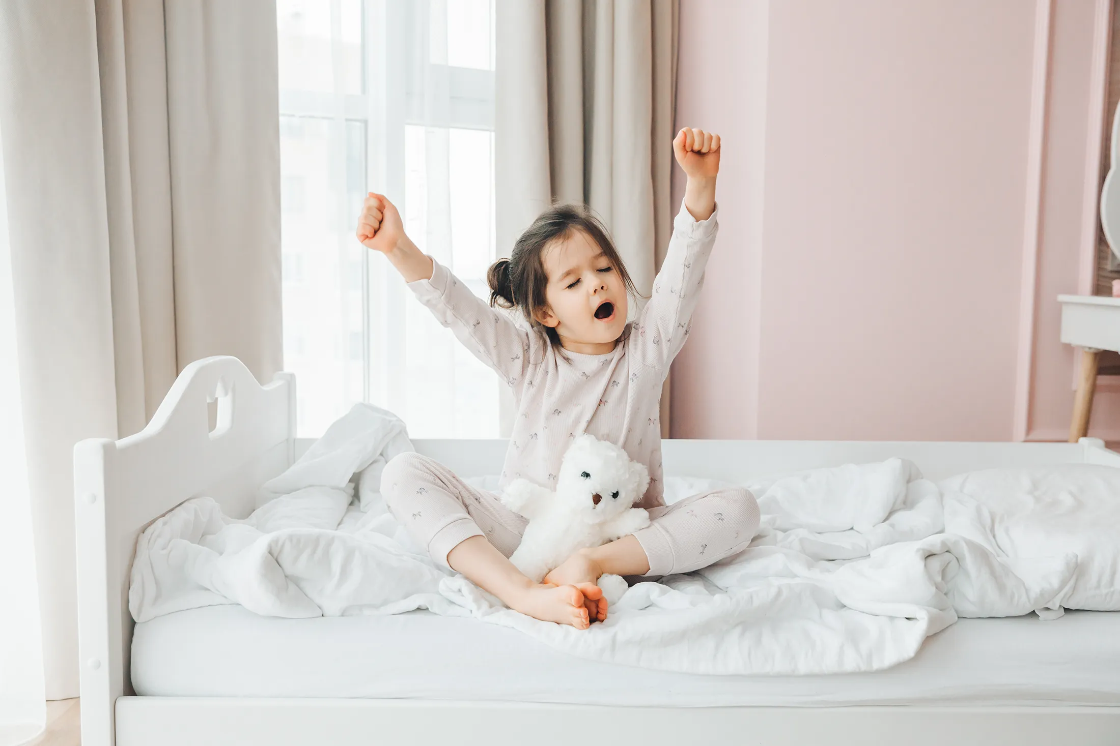 a-cute-baby-girl-enjoys-a-sunny-morning-good-morn-2025-03-05-21-47-15-utc Young girl stretching and yawning in bed during a bright morning, holding a white teddy bear and surrounded by cozy white bedding.