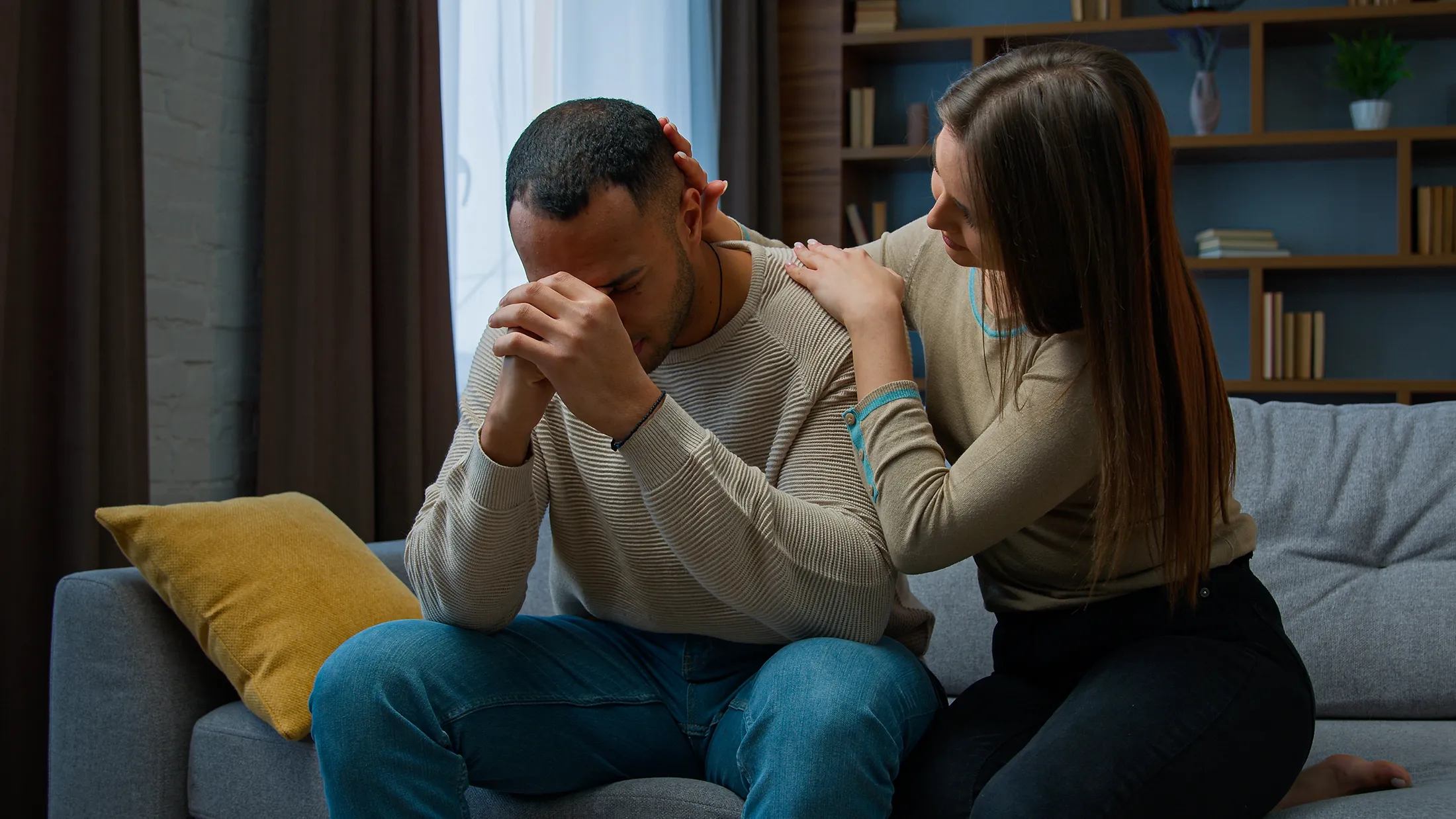 One partner comforting the other by placing a hand on their shoulder while they sit on a couch, illustrating emotional support during relationship stress or anxiety.