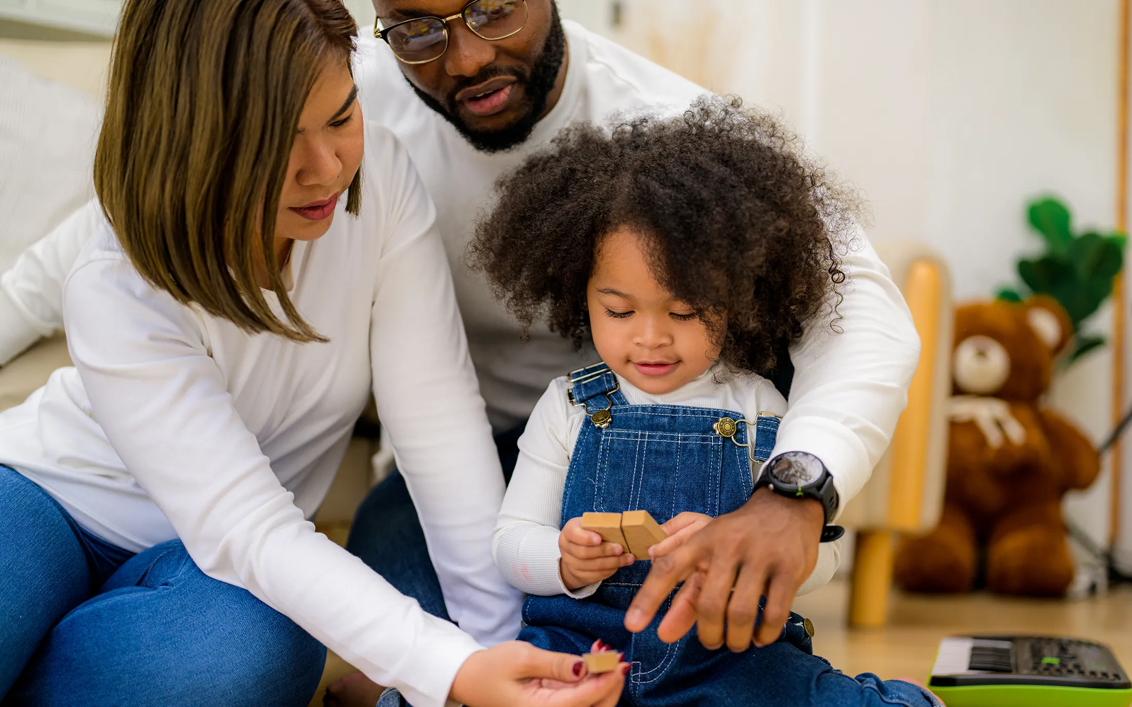 Parents sitting with their young daughter, helping her play with wooden blocks, smiling and engaged in a cozy home environment.